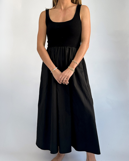 A woman stands against a plain light background, wearing a black sleeveless dress. Her arms are crossed lightly in front, showcasing her bracelets.