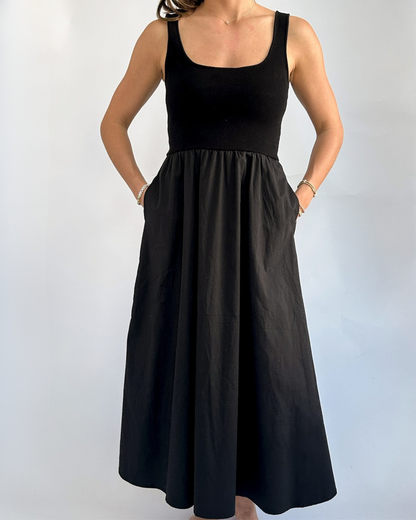 Woman in sleeveless black dress with a fitted bodice and flowing skirt stands against a light background, hands in pockets, conveying a relaxed vibe.