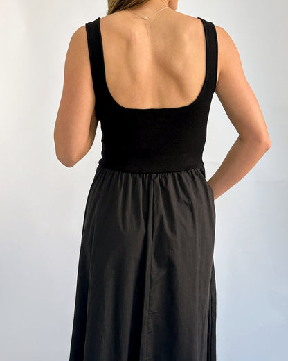 Woman in sleeveless black dress with a fitted bodice and flowing skirt stands against a light background, hand in pocket, conveying a relaxed vibe.