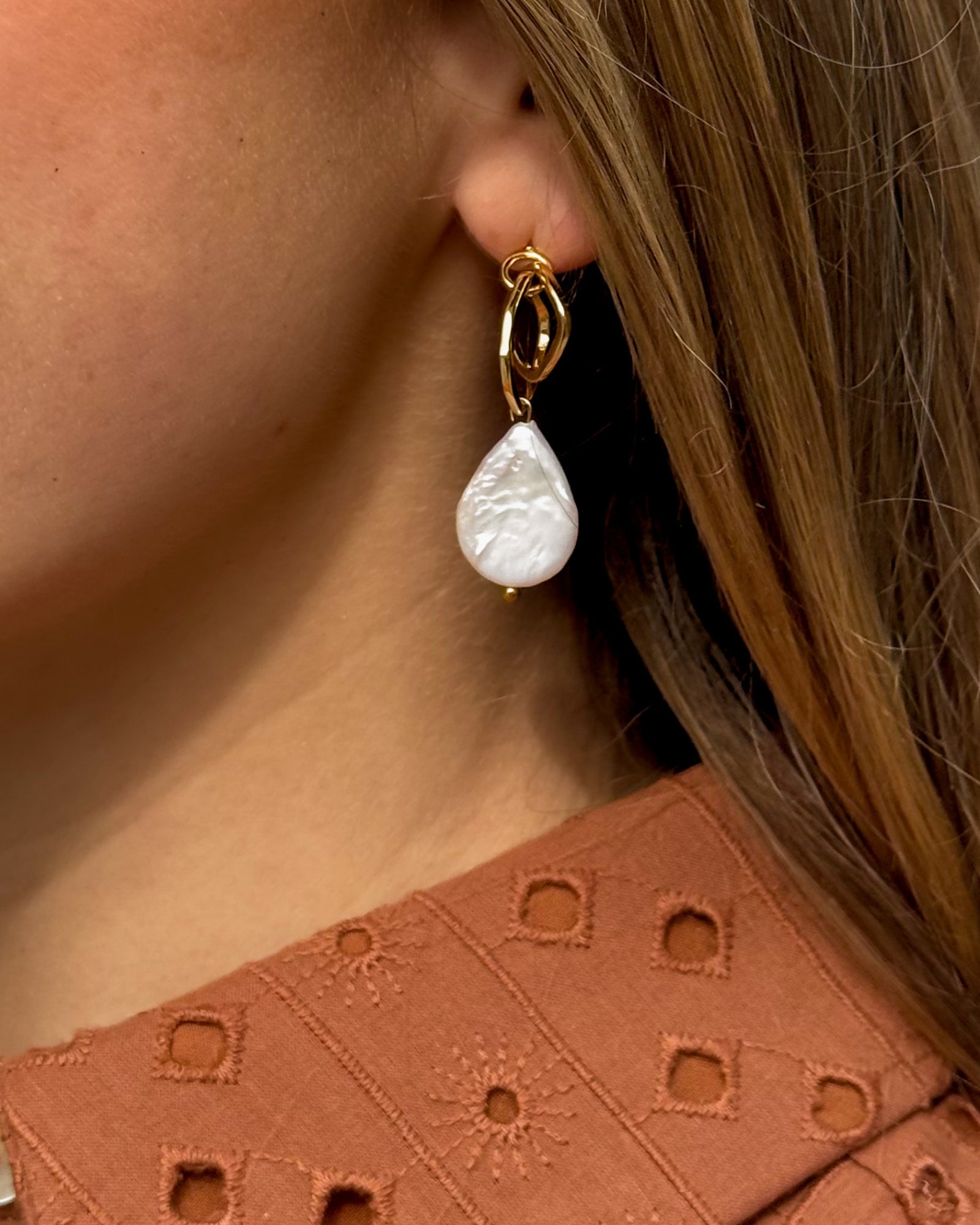 Close-up of a woman's ear showcasing a gold earring with a teardrop-shaped white pearl. She wears a textured, rust-colored blouse. Elegant and stylish.