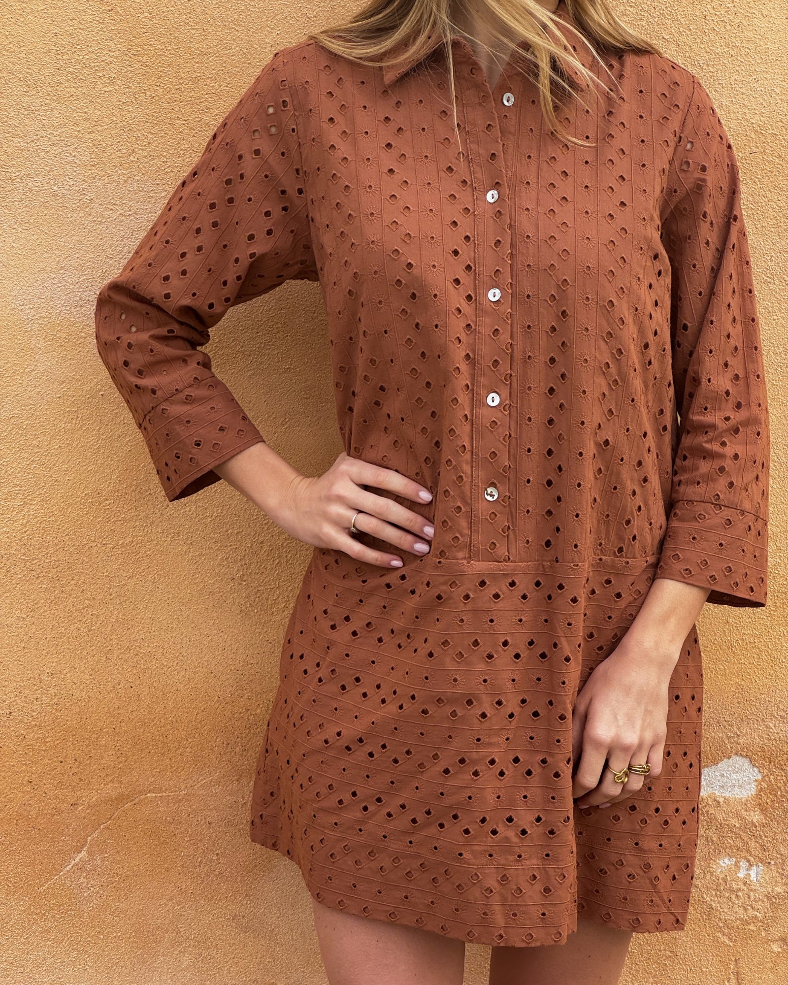 A woman stands against a textured, tan wall wearing a rust-colored, eyelet-patterned dress with white buttons. Her hand rests on her hip, conveying a relaxed vibe.