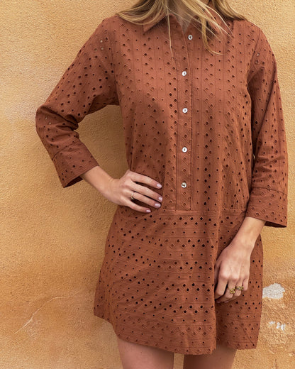 A woman stands against a textured, tan wall wearing a rust-colored, eyelet-patterned dress with white buttons. Her hand rests on her hip, conveying a relaxed vibe.