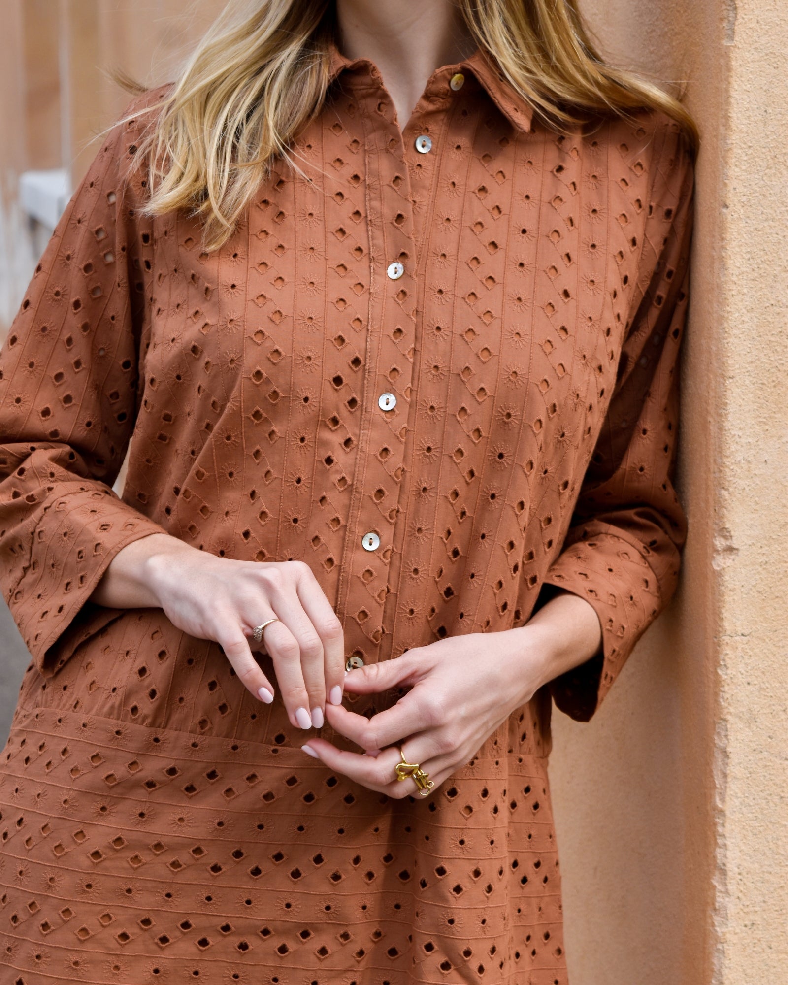 A woman stands against a textured, tan wall wearing a rust-colored, eyelet-patterned dress with white buttons.