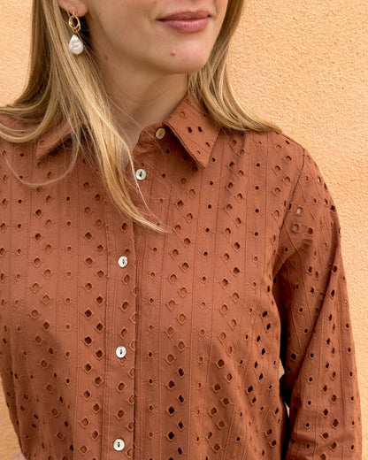 A woman stands against a textured, tan wall wearing a rust-colored, eyelet-patterned dress with white buttons. 