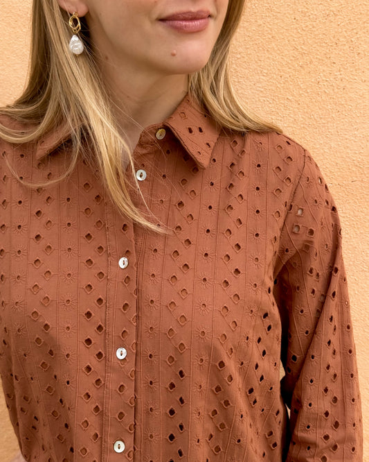 A woman stands against a textured, tan wall wearing a rust-colored, eyelet-patterned dress with white buttons. 