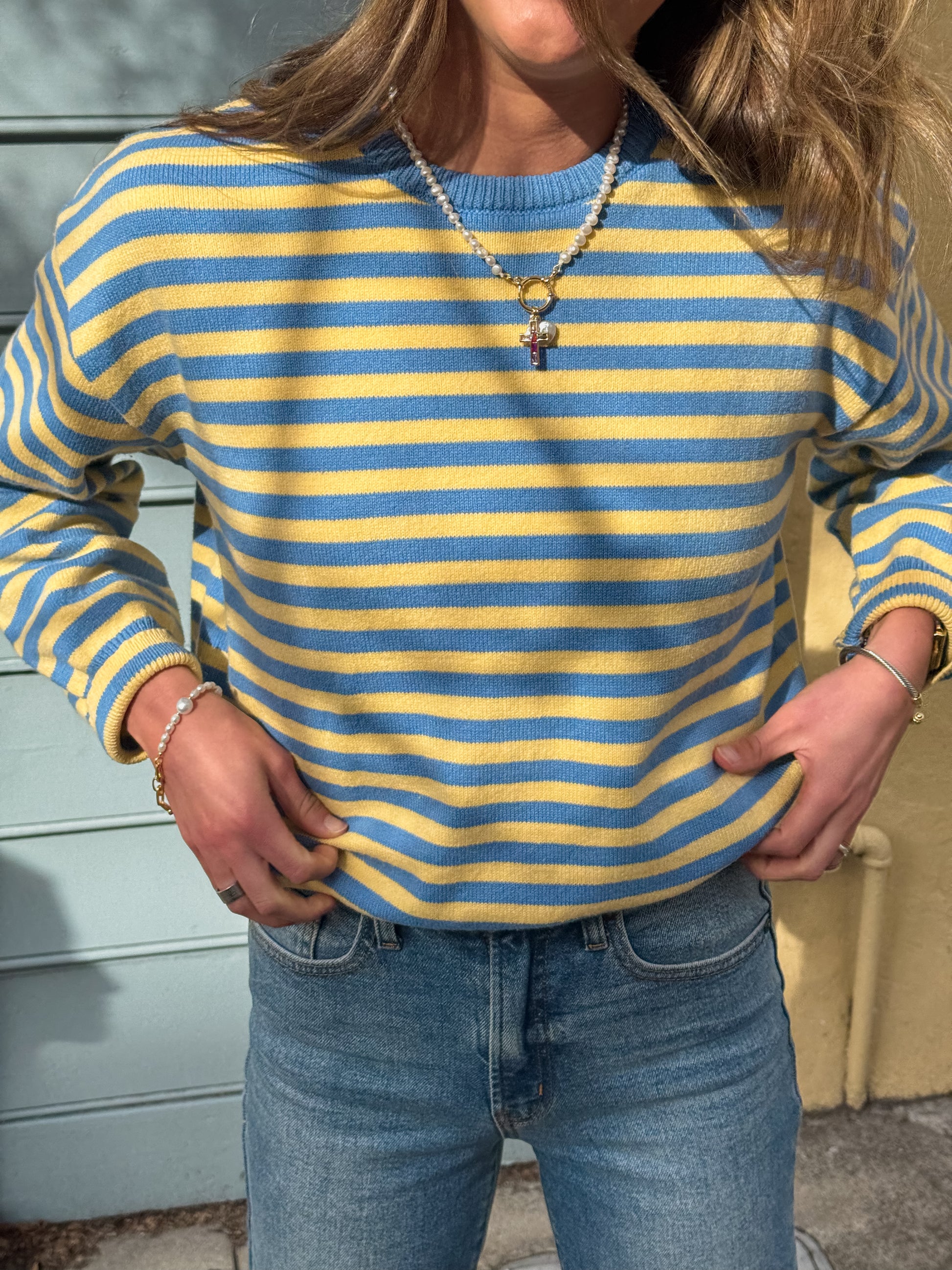 Person in a blue and yellow striped sweater and jeans stands in sunlight, hands in pockets. Wavy hair and layered necklaces complete the casual look.