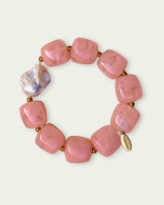 A bracelet featuring smooth square pink beads and one irregular pearl-like bead, linked by small gold spheres. An engraved gold tag is attached.