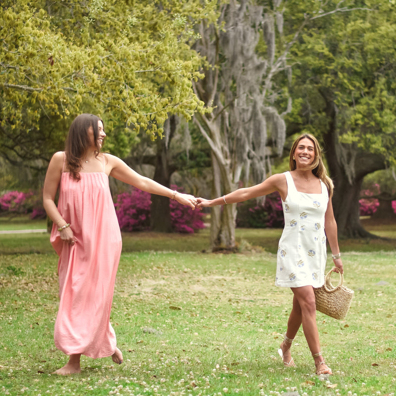 Two women holding hands and walking in a lush green park. One wears a white dress, the other a pink dress. They appear joyful and relaxed.