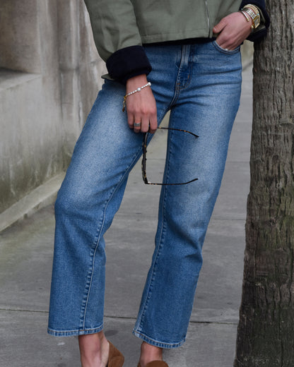 A person in blue jeans and a green jacket leans casually against a tree. They're holding sunglasses and wearing several bracelets, creating a relaxed vibe.