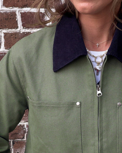Person in a green jacket with a black collar over a navy sweater and white shirt, wearing blue jeans and necklaces, stands against a brick wall.