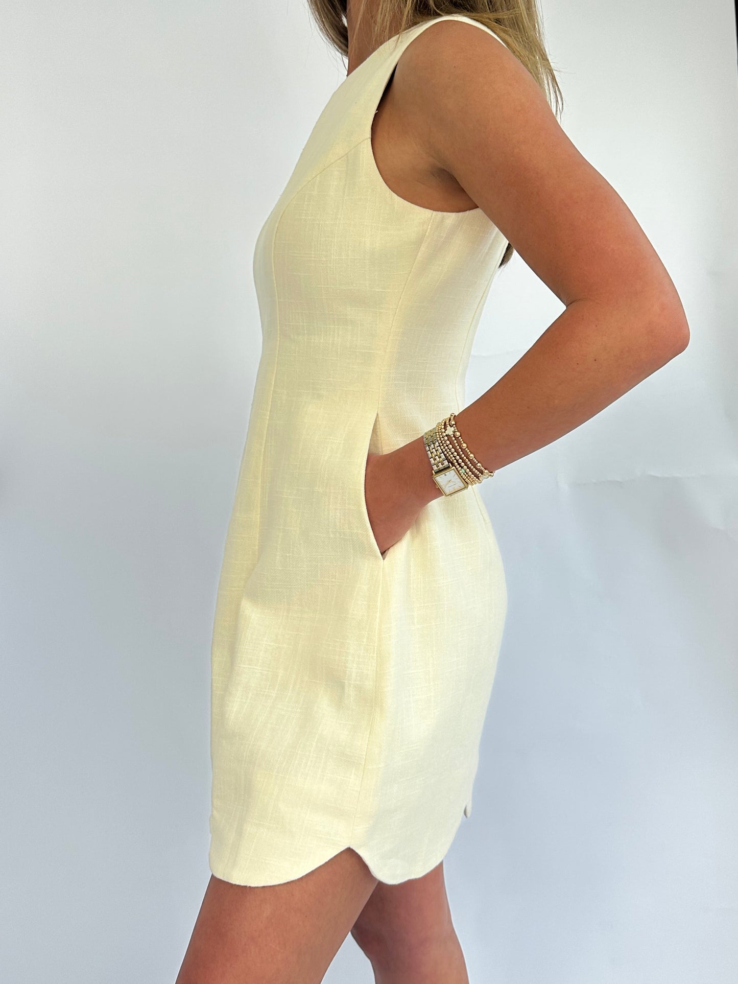 A person wearing a sleeveless, light yellow dress with a scalloped hem. The background is plain white, and they have a bracelet on each wrist.