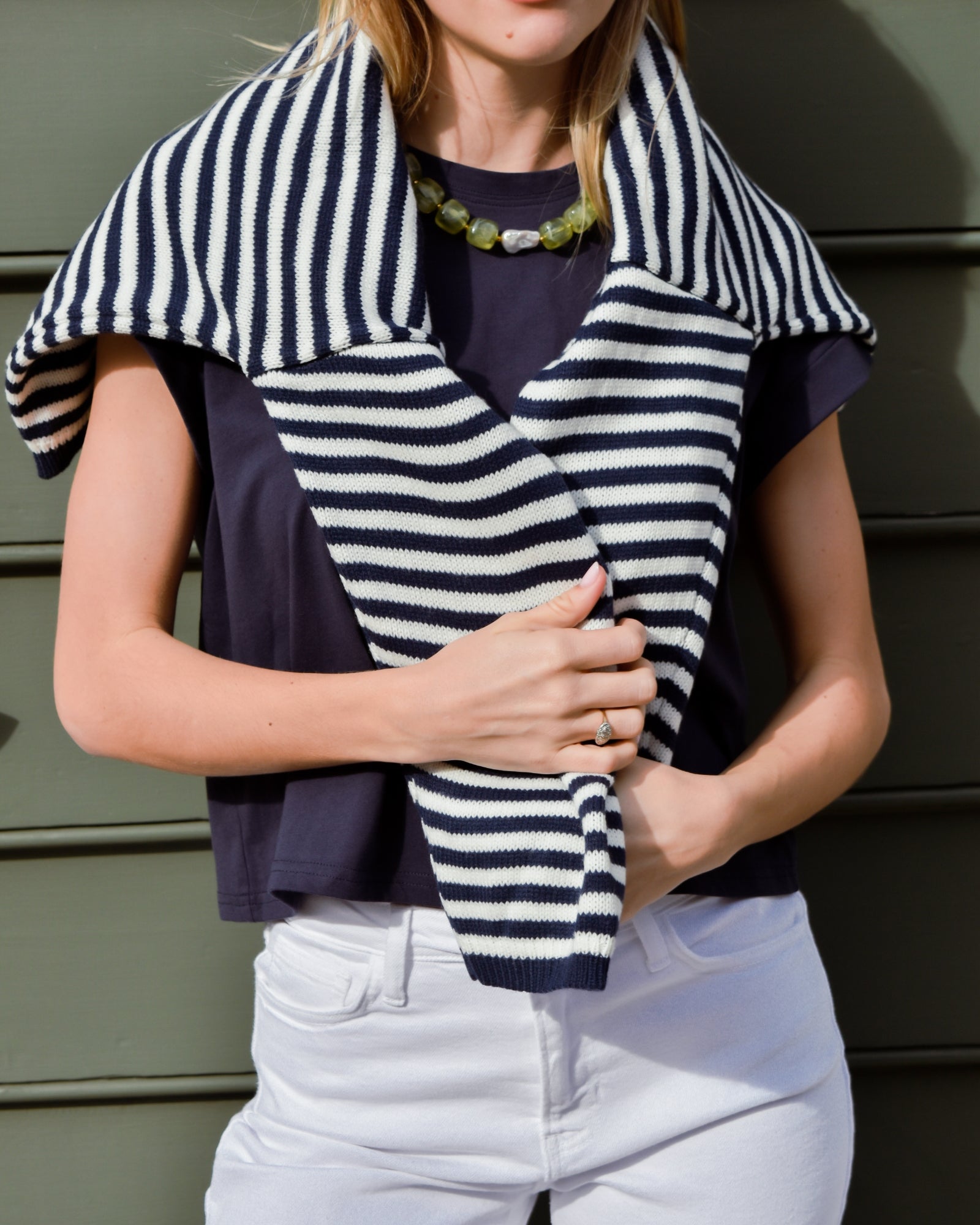 A person wearing a navy shirt and white pants drapes a black and white striped sweater over their shoulders. They hold the sweater with one hand, wearing a green beaded necklace.