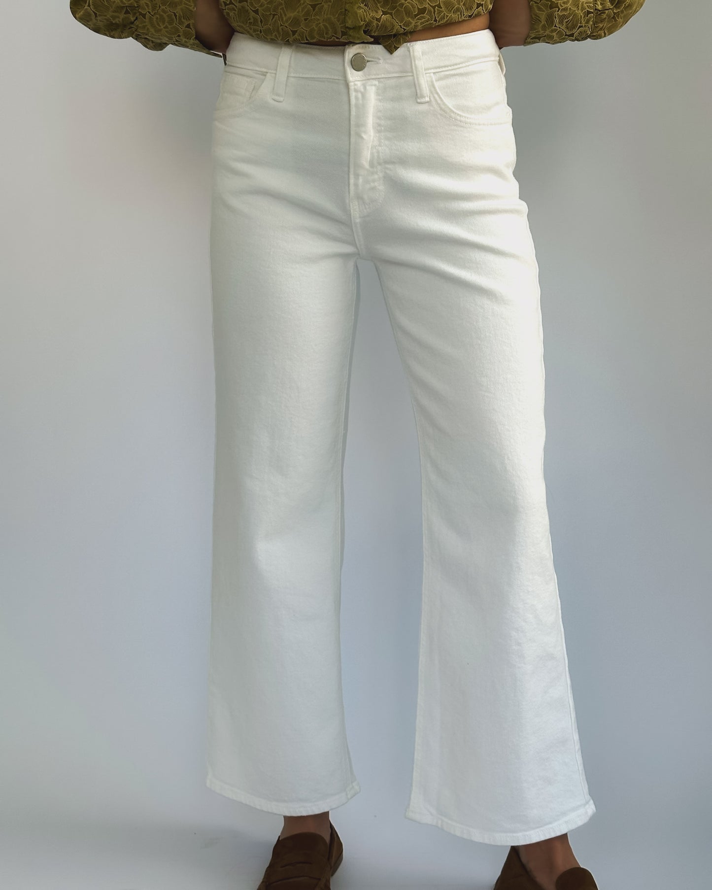 Wide-leg white jeans on a person against a plain background. They wear a green patterned top and brown shoes, creating a casual, stylish look.