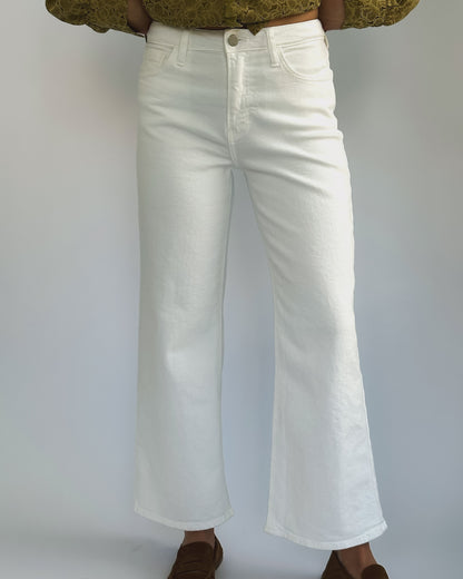 Wide-leg white jeans on a person against a plain background. They wear a green patterned top and brown shoes, creating a casual, stylish look.