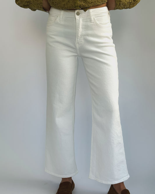 Wide-leg white jeans on a person against a plain background. They wear a green patterned top and brown shoes, creating a casual, stylish look.