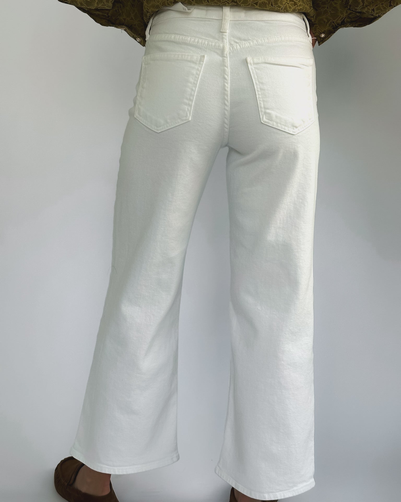 Wide-leg white jeans on a person against a plain background. They wear a green patterned top and brown shoes, creating a casual, stylish look.