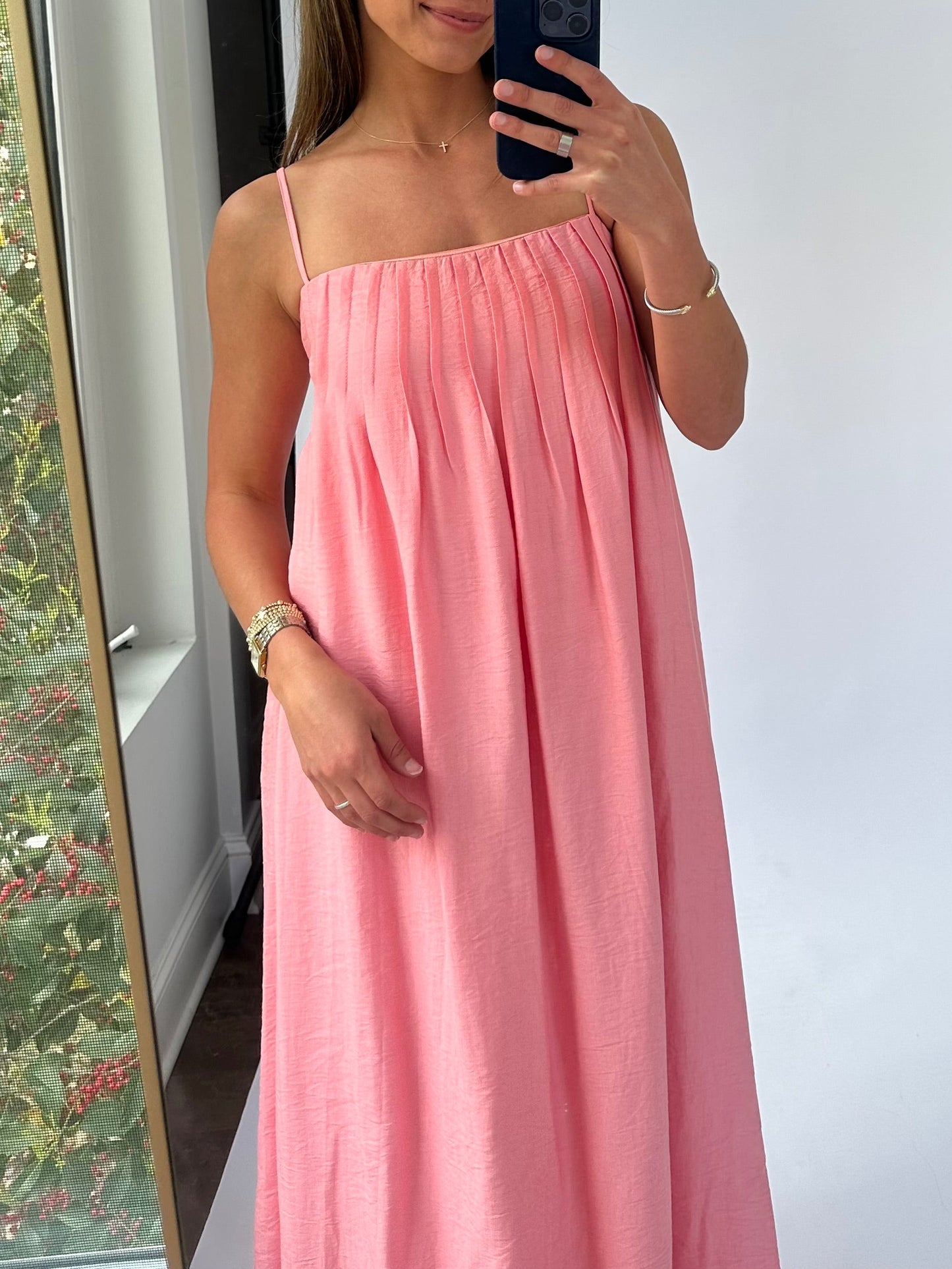 A woman wearing a flowing pink sundress with thin straps takes a mirror selfie. She smiles softly, exuding a relaxed and confident vibe.