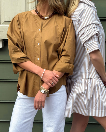 Two people stand side by side against a green background. One wears a brown button-up shirt and white pants, the other a striped dress. Both wear accessories.