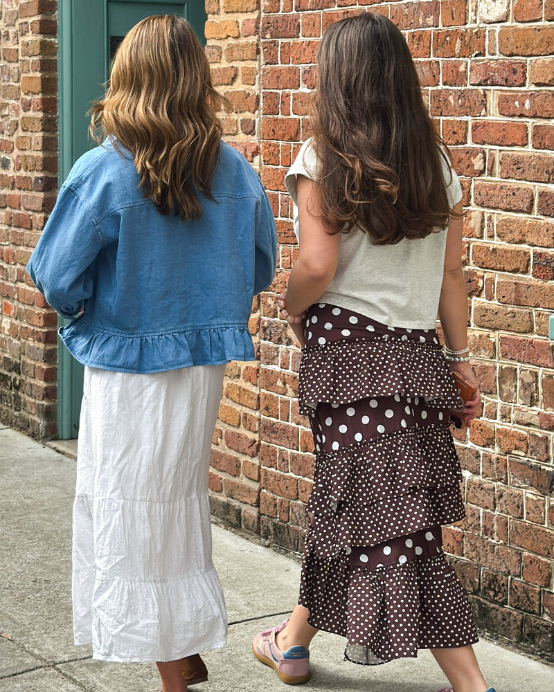 Two women walking side by side on a sidewalk. One wears a blue jacket and white skirt, the other a gray top with a polka dot skirt. Brick wall background.