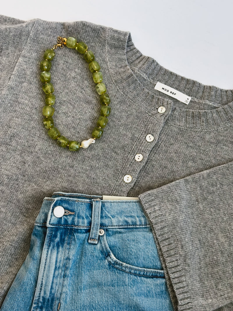 Gray knit sweater with white buttons paired with light blue denim jeans. A green beaded necklace is elegantly draped over the sweater.
