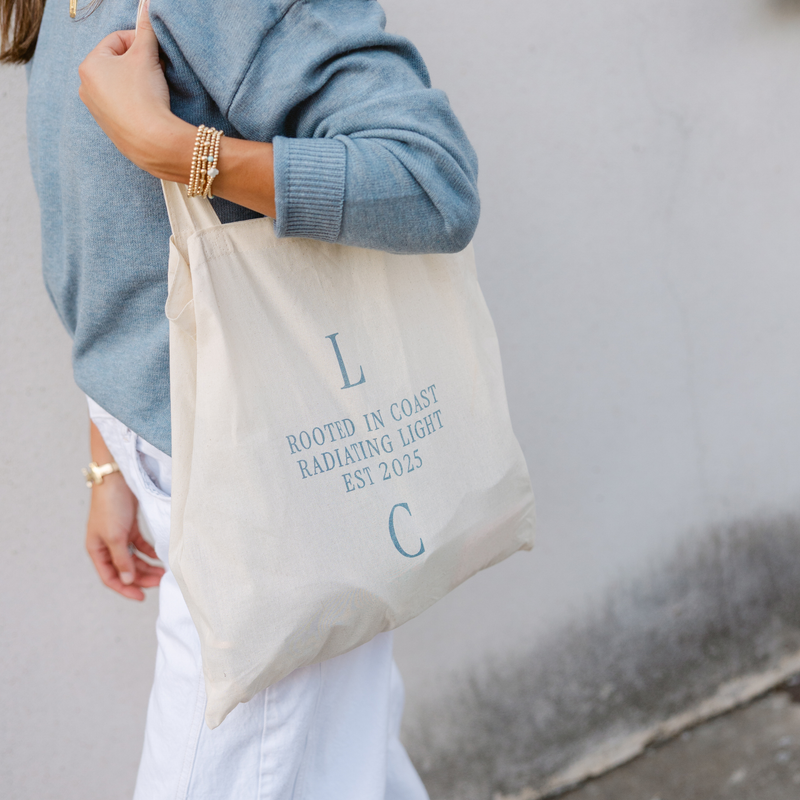 A person in a blue sweater carries a cream tote bag with blue text: "Rooted in Coast, Radiating Light, Est 2025." The mood is casual and stylish.
