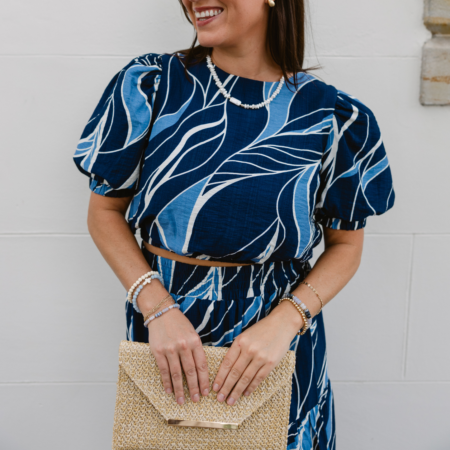 Smiling woman in a blue patterned outfit holds a straw clutch. She wears pearl jewelry, standing against a plain white wall, exuding a stylish and cheerful vibe.