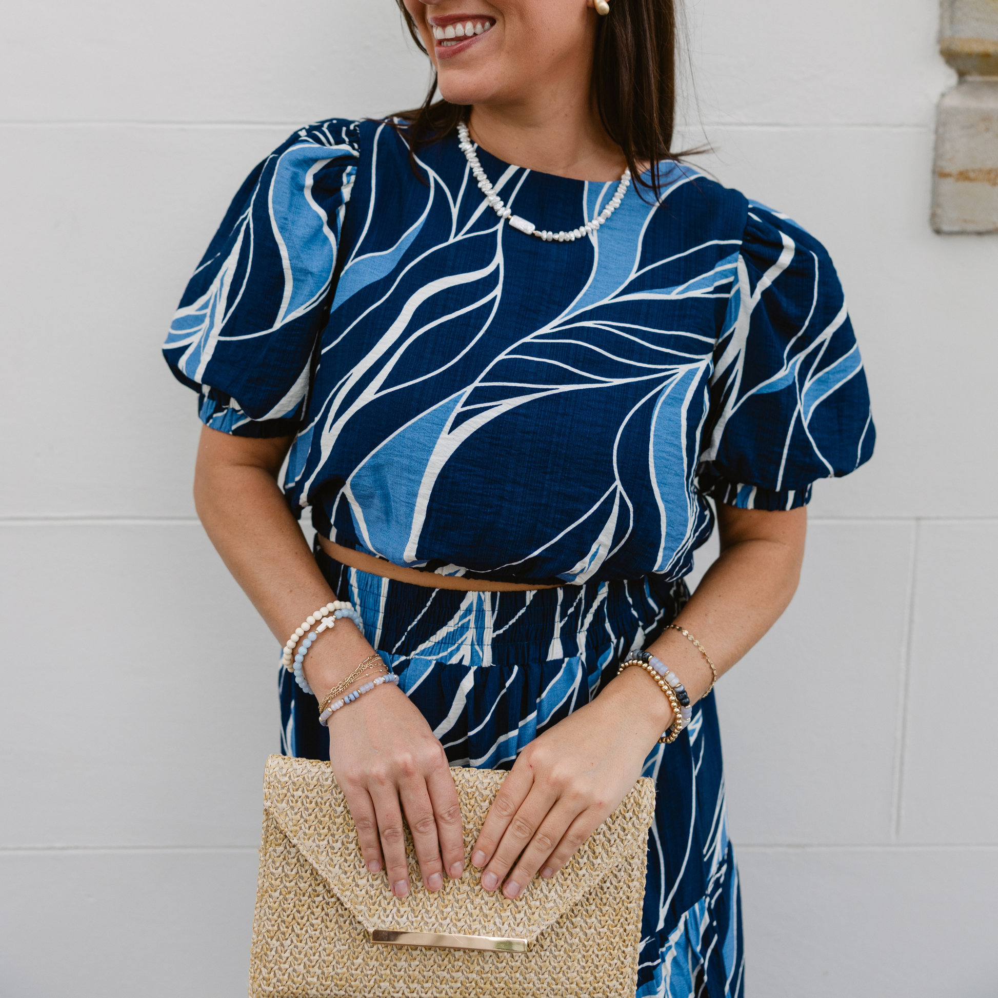 Smiling woman in a blue patterned outfit holds a straw clutch. She wears pearl jewelry, standing against a plain white wall, exuding a stylish and cheerful vibe.
