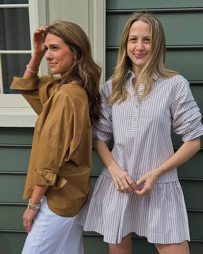 Two women stand smiling in front of a green building. One wears a brown top with white pants, the other a striped dress. The mood is friendly and relaxed.
