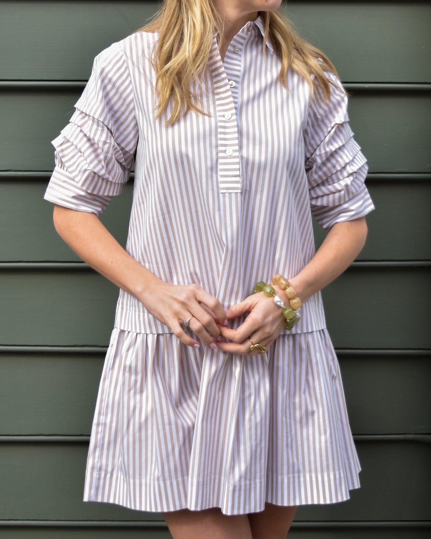 Woman in a striped shirt with puffed sleeves stands against a gray background. Her blonde hair is down, and she wears pearl earrings. The mood is casual.