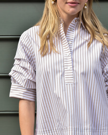 Woman in a striped shirt with puffed sleeves stands against a gray background. Her blonde hair is down, and she wears pearl earrings. The mood is casual.