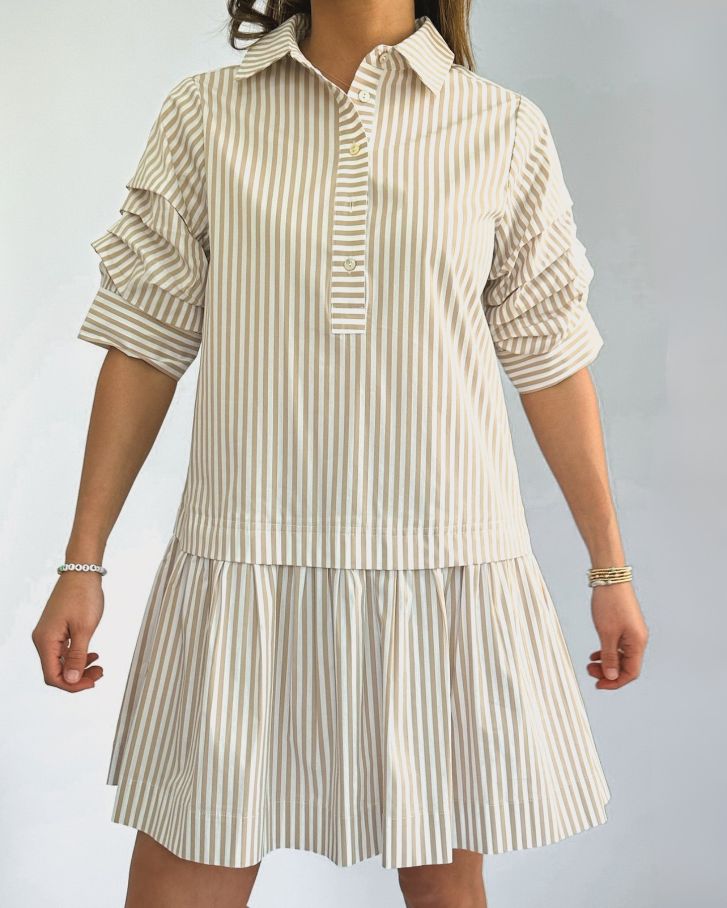 A person wearing a beige and white vertically striped shirtdress with ruffled sleeves. The dress has a collared neckline and falls above the knee.