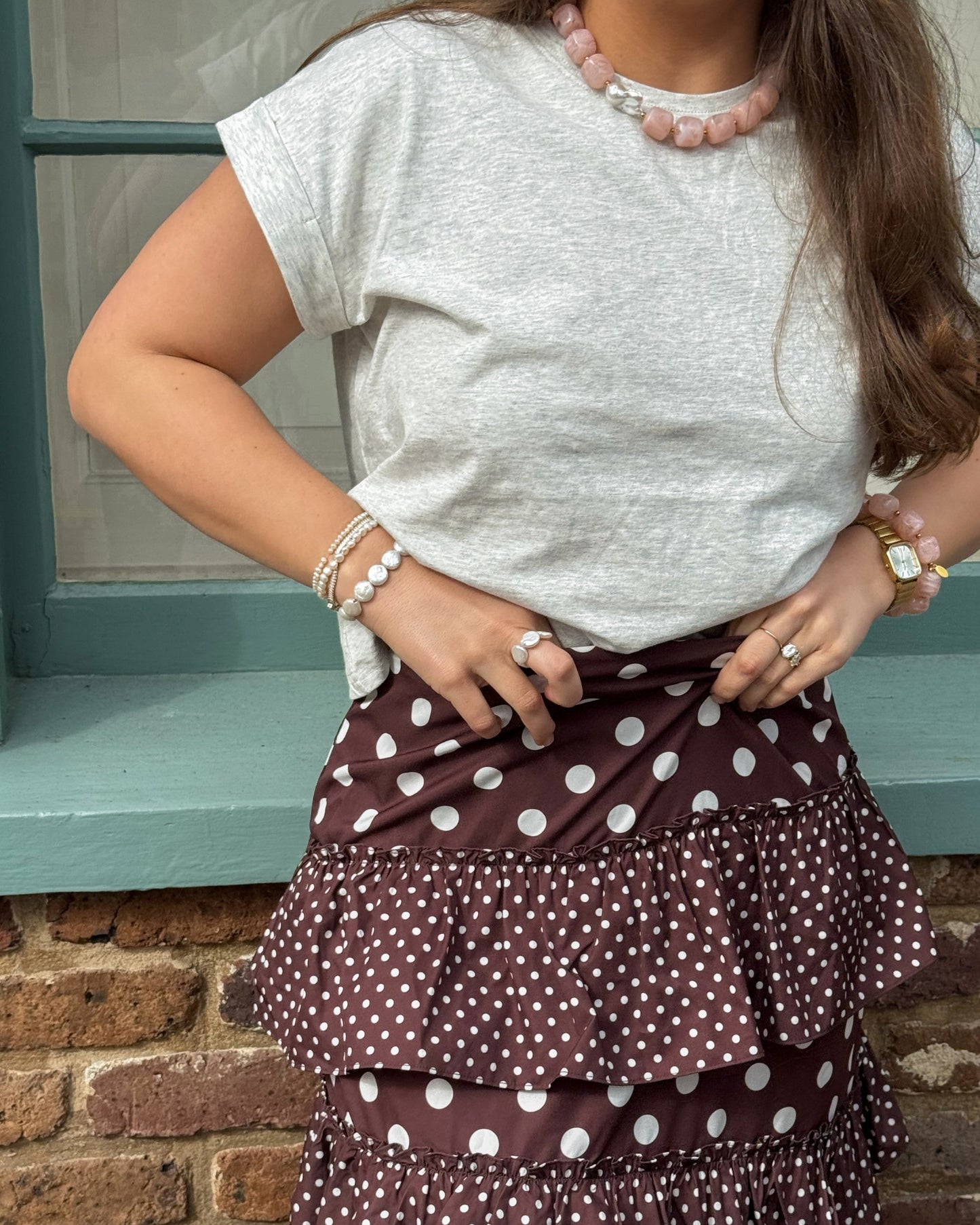 A person wearing a light gray t-shirt and a brown polka dot tiered skirt stands by a brick wall. They have a pink necklace and bracelets, conveying a casual, stylish look.