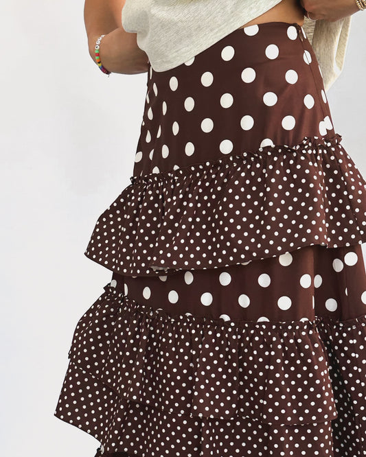 A stylish, brown layered skirt with white polka dots. The tiers vary in dot size, creating a playful and elegant look. Worn with bare feet.