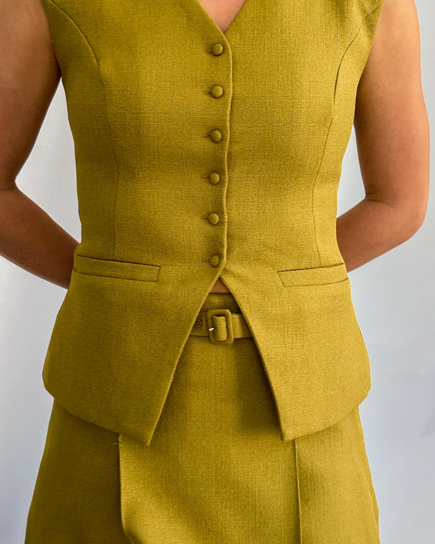 A person wearing a stylish olive sleeveless vest with buttons, paired with a matching skirt. Their arms are crossed behind their back.