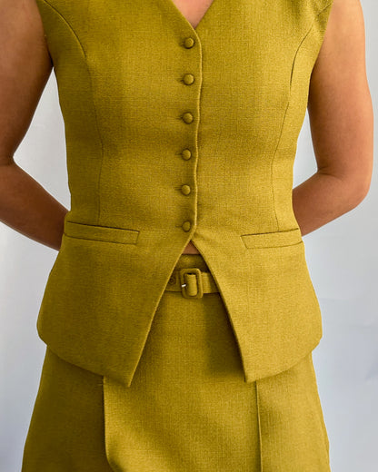 A person wearing a stylish olive sleeveless vest with buttons, paired with a matching skirt. Their arms are crossed behind their back.
