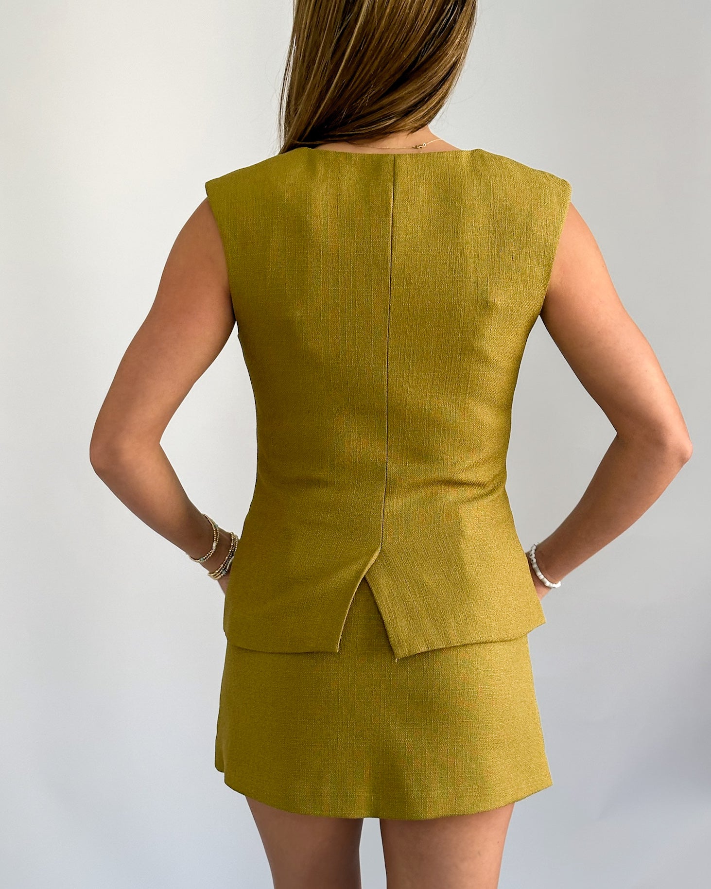 A person wearing a stylish olive sleeveless vest with buttons, paired with a matching skirt. 