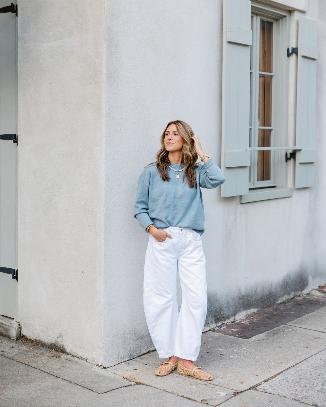 A woman in a blue sweater and white pants stands by a light-colored wall with a window. She looks content, with one hand in her pocket and the other touching her hair.