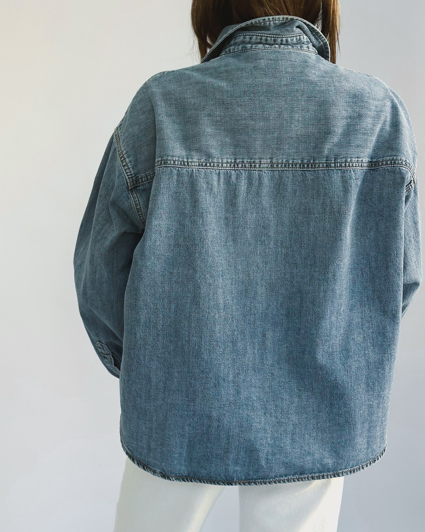 A person wearing a loose-fitting denim jacket, layered over a light-colored t-shirt and paired with white pants. The casual style suggests comfort and ease.