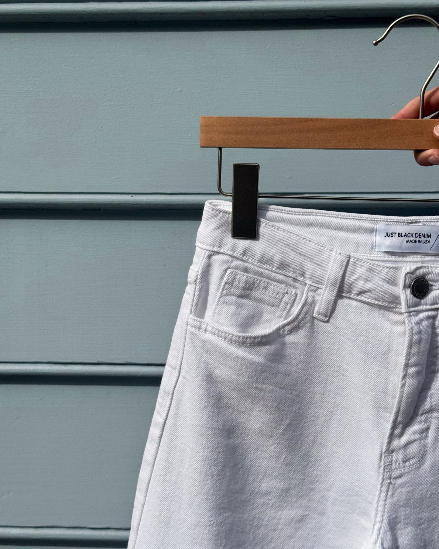 White denim jeans on a wooden hanger against a blue wall. A label reads "Just Black Denim, Made in USA." The scene feels minimal and modern.