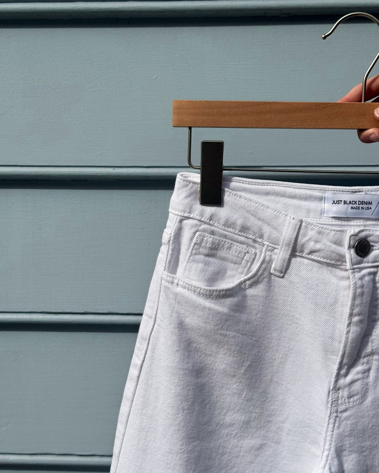 White denim jeans on a wooden hanger against a blue wall. A label reads "Just Black Denim, Made in USA." The scene feels minimal and modern.